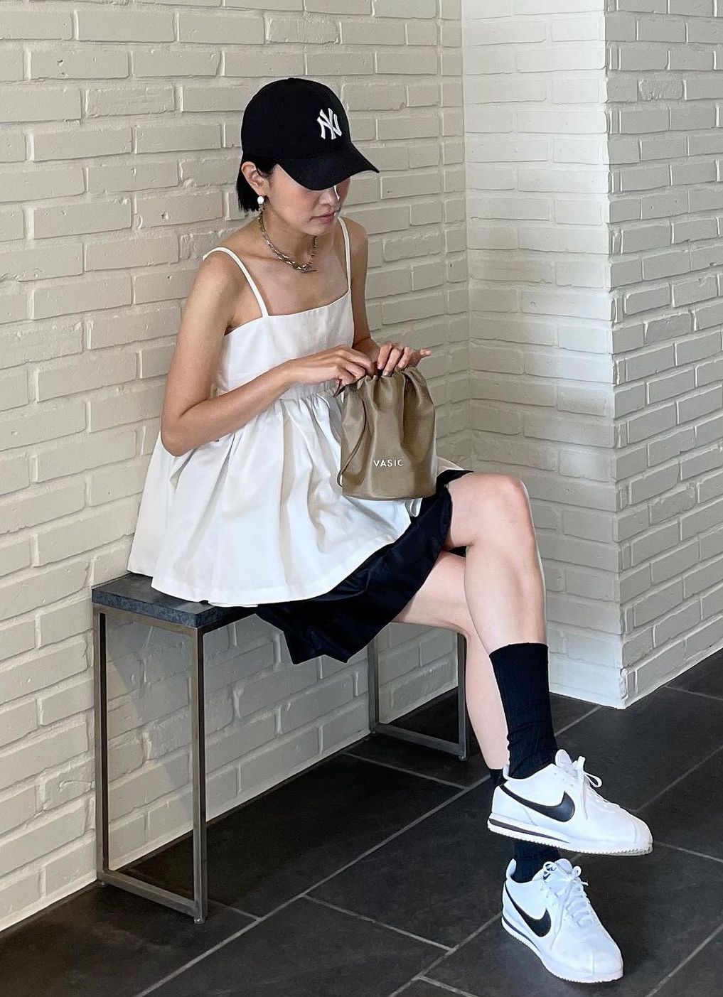 A woman sitting on a bench wearing a white babydoll camisole, black mini skirt, black Yankees baseball cap, and white Nike sneakers with black socks.