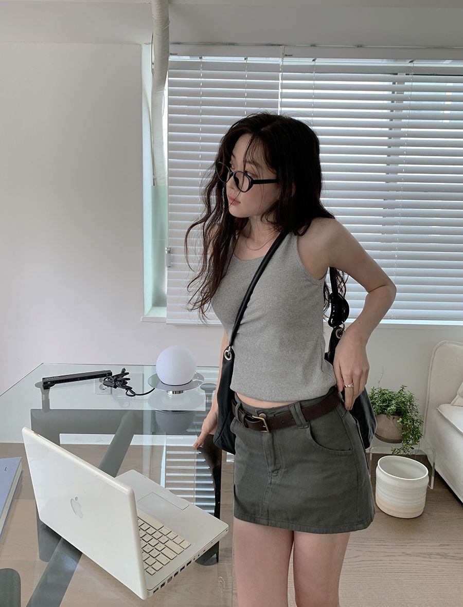 A woman with long wavy hair and glasses wearing a grey ribbed tank top and an olive green denim mini skirt with a brown belt, standing in a bright modern room.