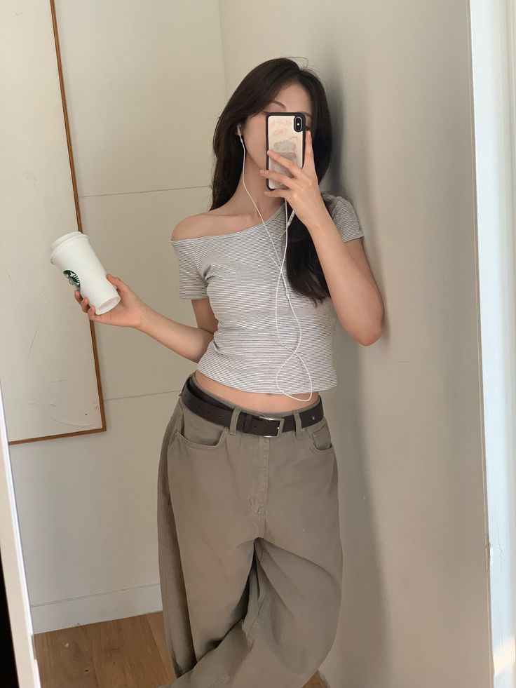 A woman taking a mirror selfie wearing a grey and white striped one-shoulder t-shirt, loose khaki trousers with a brown belt, and white wired earphones.