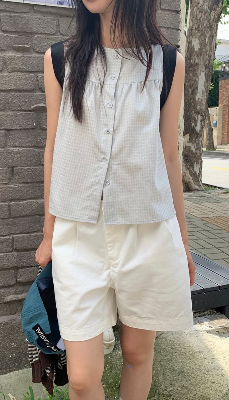 A woman standing outdoors wearing a light blue and white gingham sleeveless button-up shirt paired with white pleated shorts, holding a teal baseball cap and a striped bag.