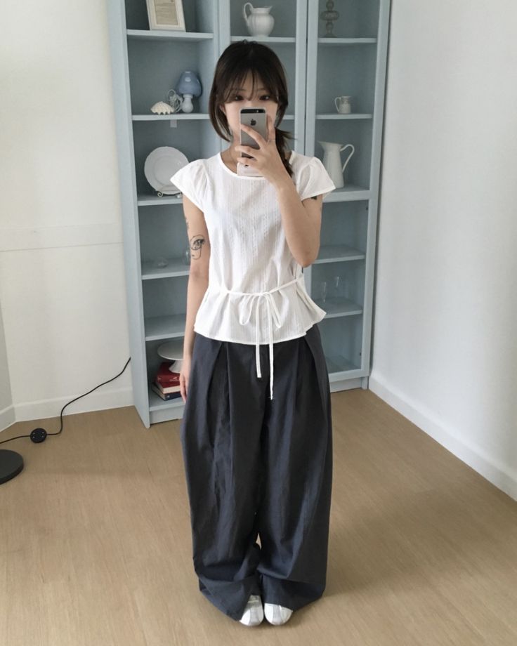 A woman taking a mirror selfie wearing a white short-sleeved blouse with a drawstring waist and very baggy dark grey pleated trousers.