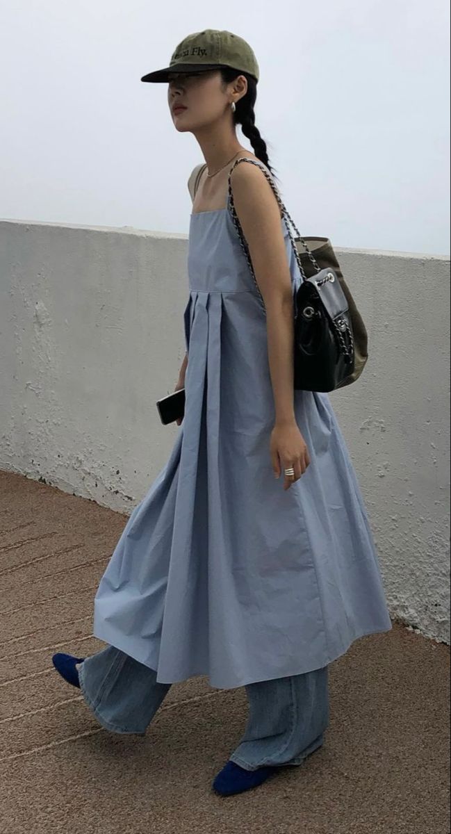 A woman walking outdoors wearing a light blue pleated sleeveless maxi dress layered over wide-leg denim jeans, styled with an olive green cap and a black shoulder bag.