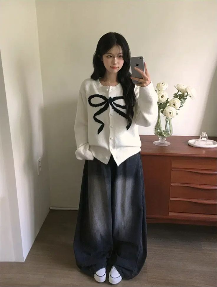 A student taking a mirror selfie wearing a cream cardigan with a large black bow design, oversized faded dark grey wide-leg jeans, and white sneakers.