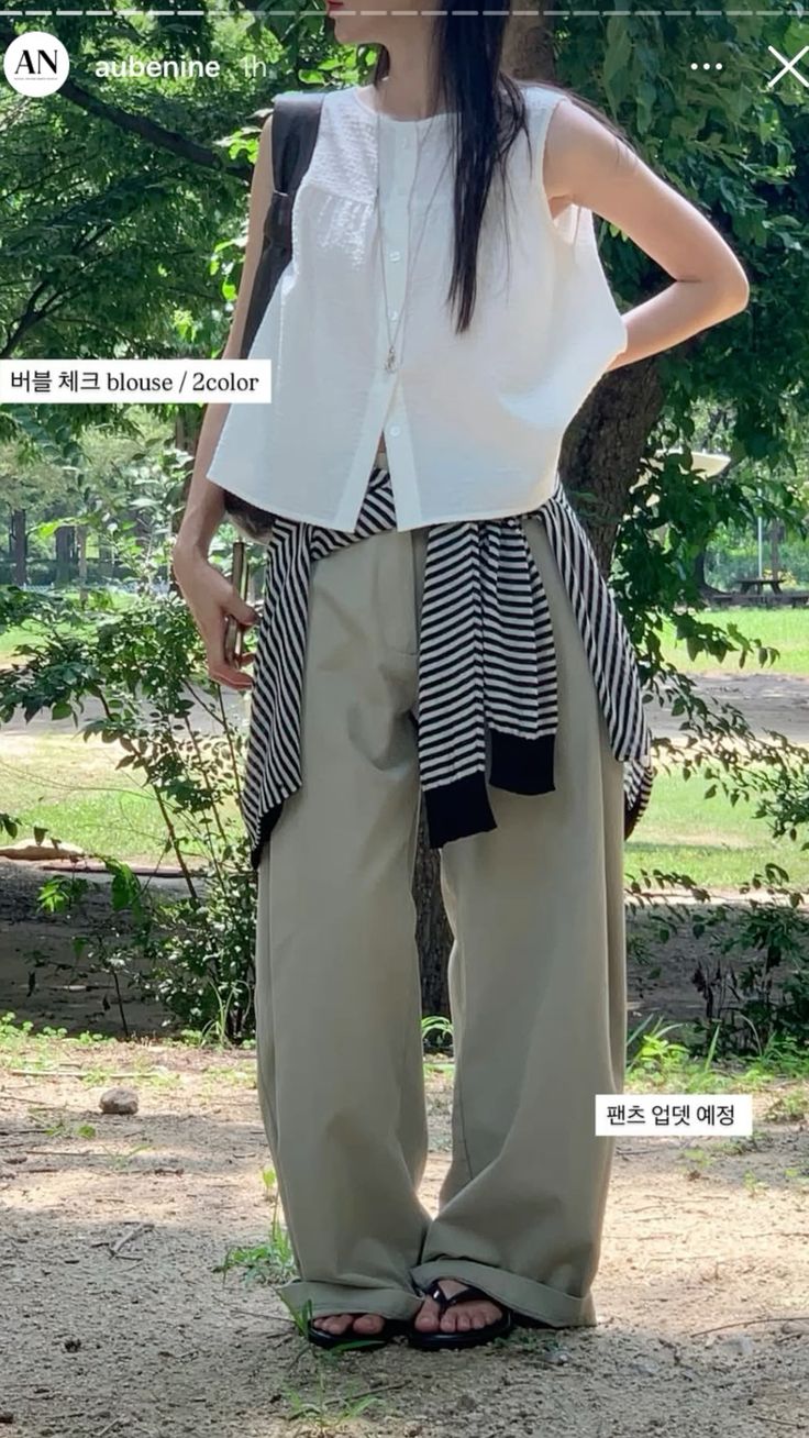 A woman standing in a park wearing a white sleeveless button-up blouse, oversized khaki trousers, and a black and white striped sweater tied around her waist.