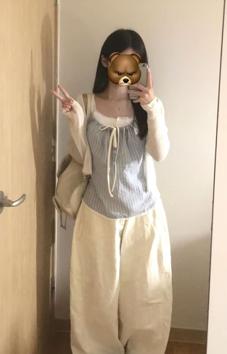 A student taking a mirror selfie wearing a blue striped camisole with a white bow, a cream cardigan, and oversized cream balloon pants with a backpack.
