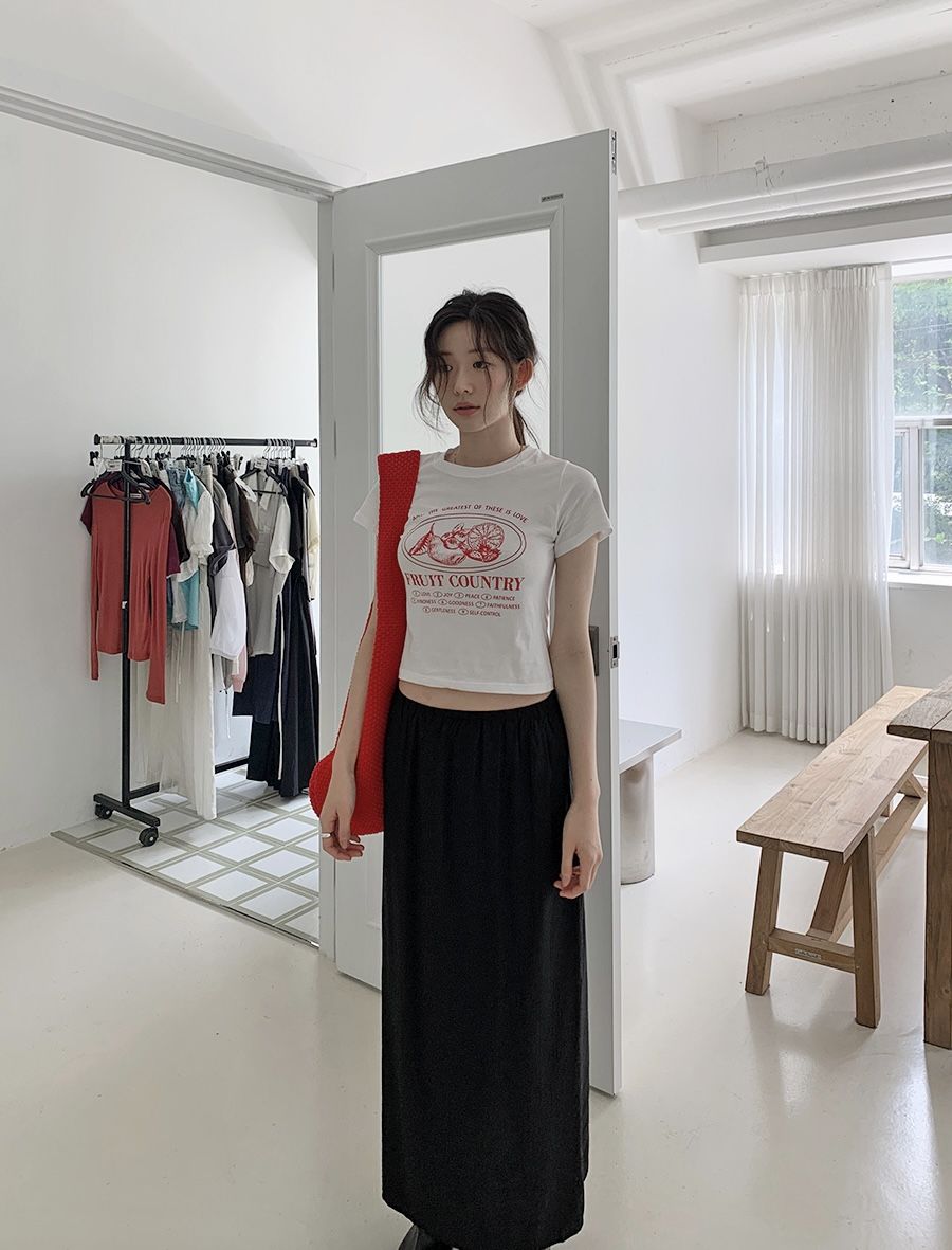 A woman standing in a white room wearing a fitted white graphic t-shirt with red text, a long black maxi skirt, and a bright red textured shoulder bag.