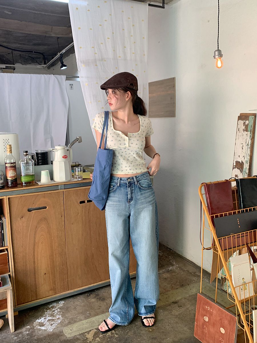 A woman standing in a rustic cafe wearing a cream floral short-sleeve henley shirt, wide-leg blue jeans, a brown flat cap, and carrying a blue tote bag.