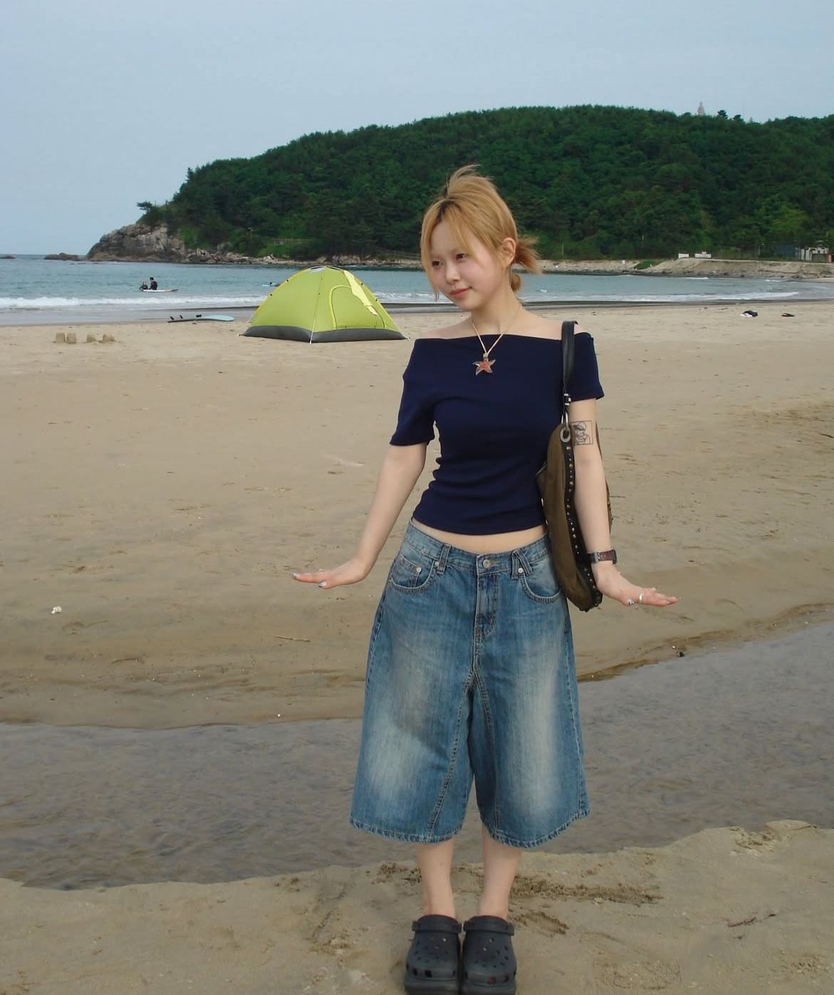 A woman standing on a beach wearing a navy blue off-the-shoulder t-shirt, baggy denim bermuda shorts, black clogs, and a large starfish pendant necklace.
