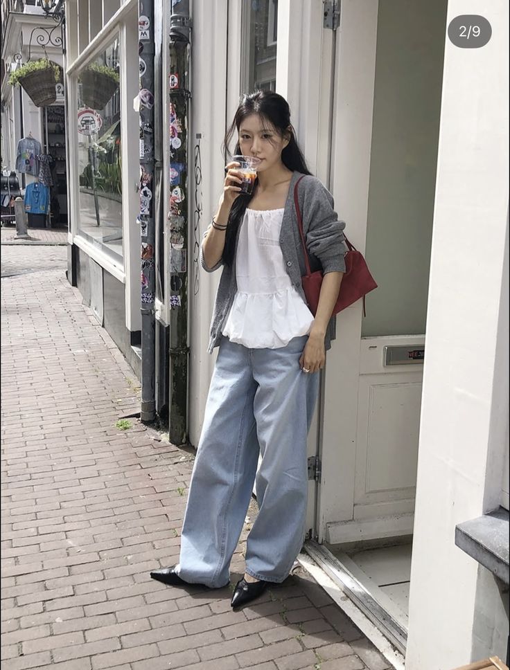 A woman is wearing a white ruffled top, light wash wide leg jeans, a gray cardigan, and black heeled shoes, holding a drink.