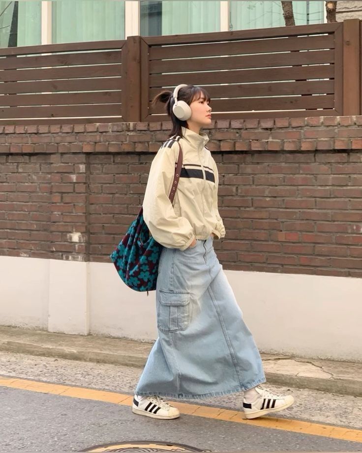 A windbreaker jacket, maxi denim skirt, and sneakers, styled with headphones and a patterned bag.
