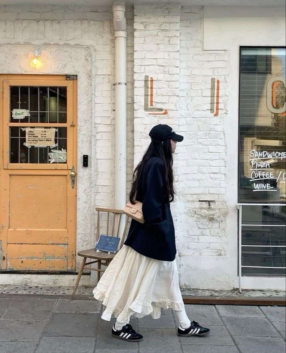 A woman wearing an oversized navy blazer, a ruffled cream skirt, white socks, and black sneakers while carrying a small pink handbag.