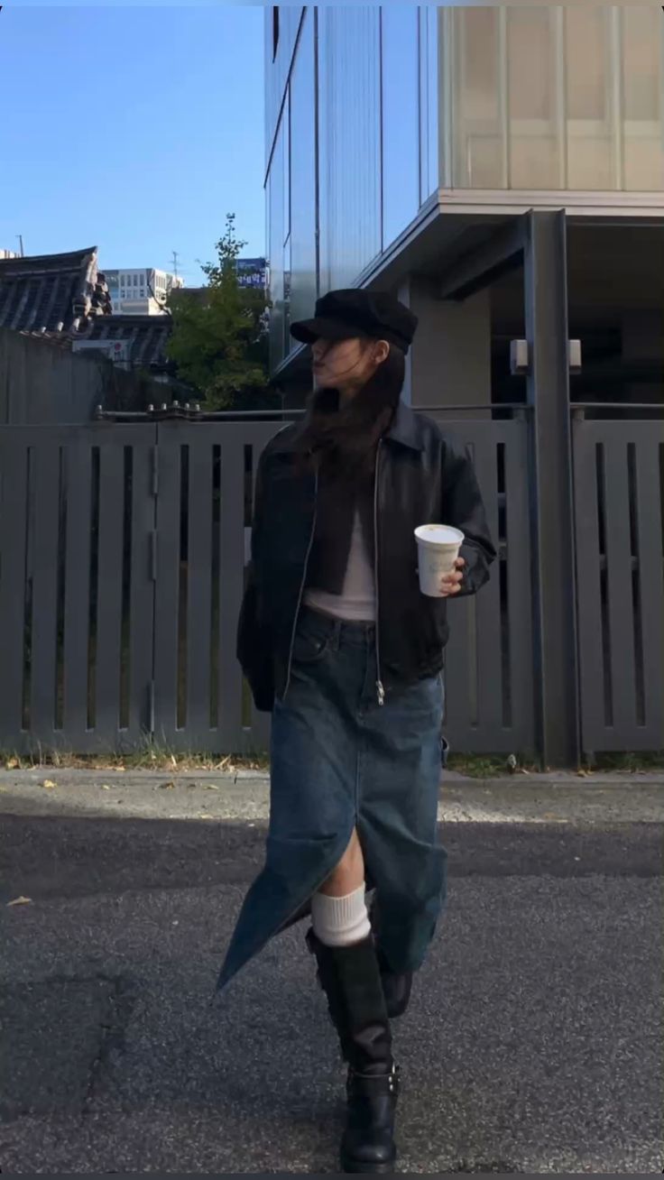 A woman wearing a black bomber jacket, a fitted top, a denim skirt, knee-high black boots, and a cap, holding a coffee cup.