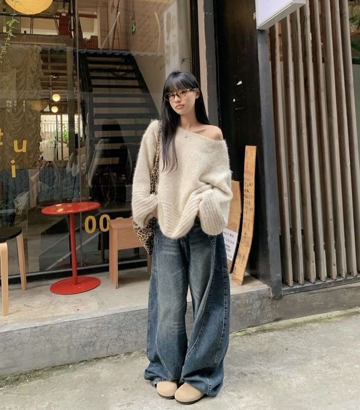 The outfit features an oversized cream knit sweater, wide leg blue jeans, and beige slip-on shoes.