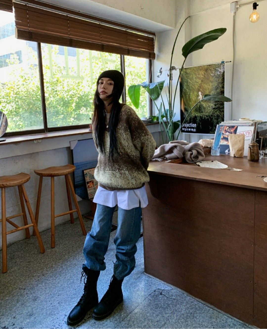 The outfit features an oversized knit sweater in brown and cream, layered over a white shirt, paired with baggy blue jeans and black lace-up boots.