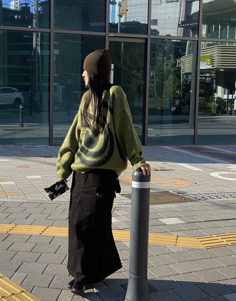 The outfit features an oversized green knit sweater with a swirl design and a black maxi skirt.