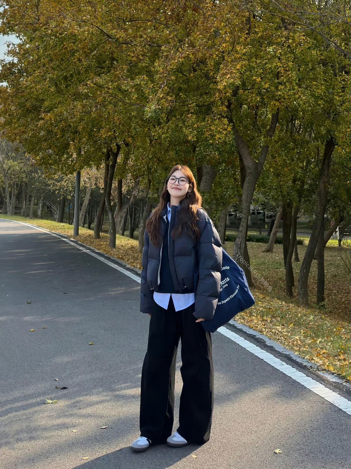 A woman wearing a gray puffer jacket, a white shirt, and black wide leg trousers with white sneakers and a blue tote bag.
