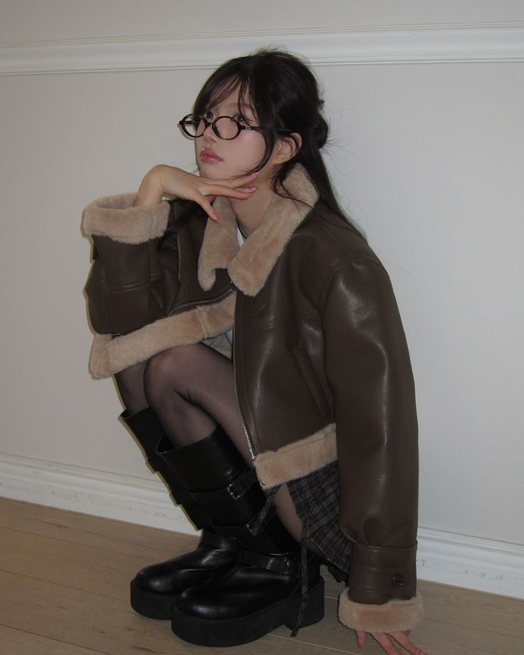 The outfit features a brown shearling jacket, a plaid skirt, black knee-high boots, and black glasses.