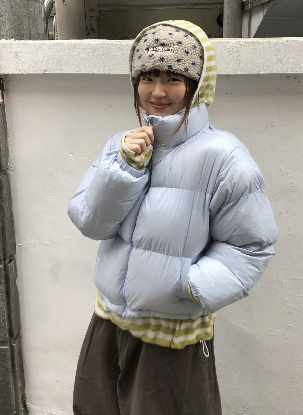 The outfit features a light blue puffer jacket layered over a striped sweater, paired with brown wide leg pants.