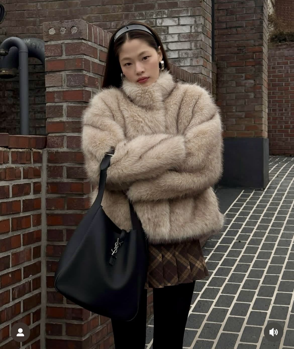 A woman is wearing a beige faux fur jacket, a plaid mini skirt, and black tights, with a black handbag.