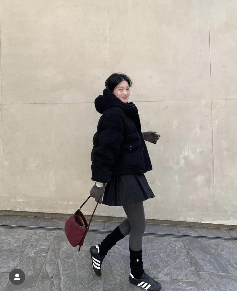 The outfit features a black puffer jacket, a black pleated mini skirt, gray tights, black sports shoes, and a burgundy handbag.