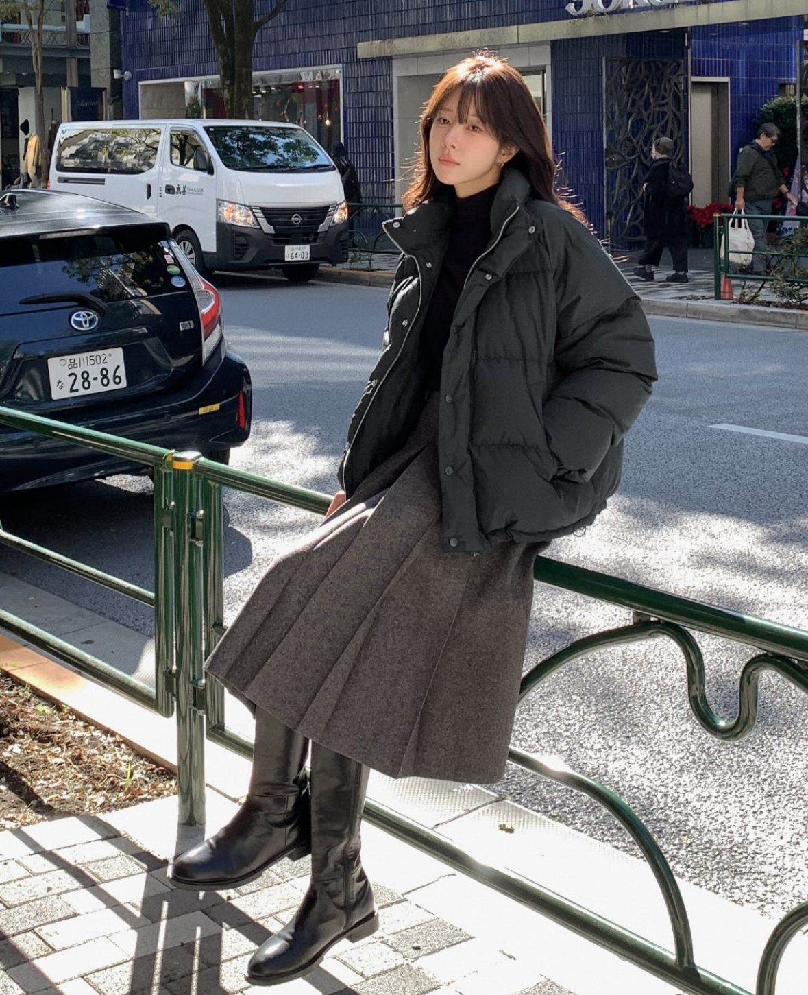 A model wearing a black puffer jacket, a gray pleated skirt, and knee-high black boots.