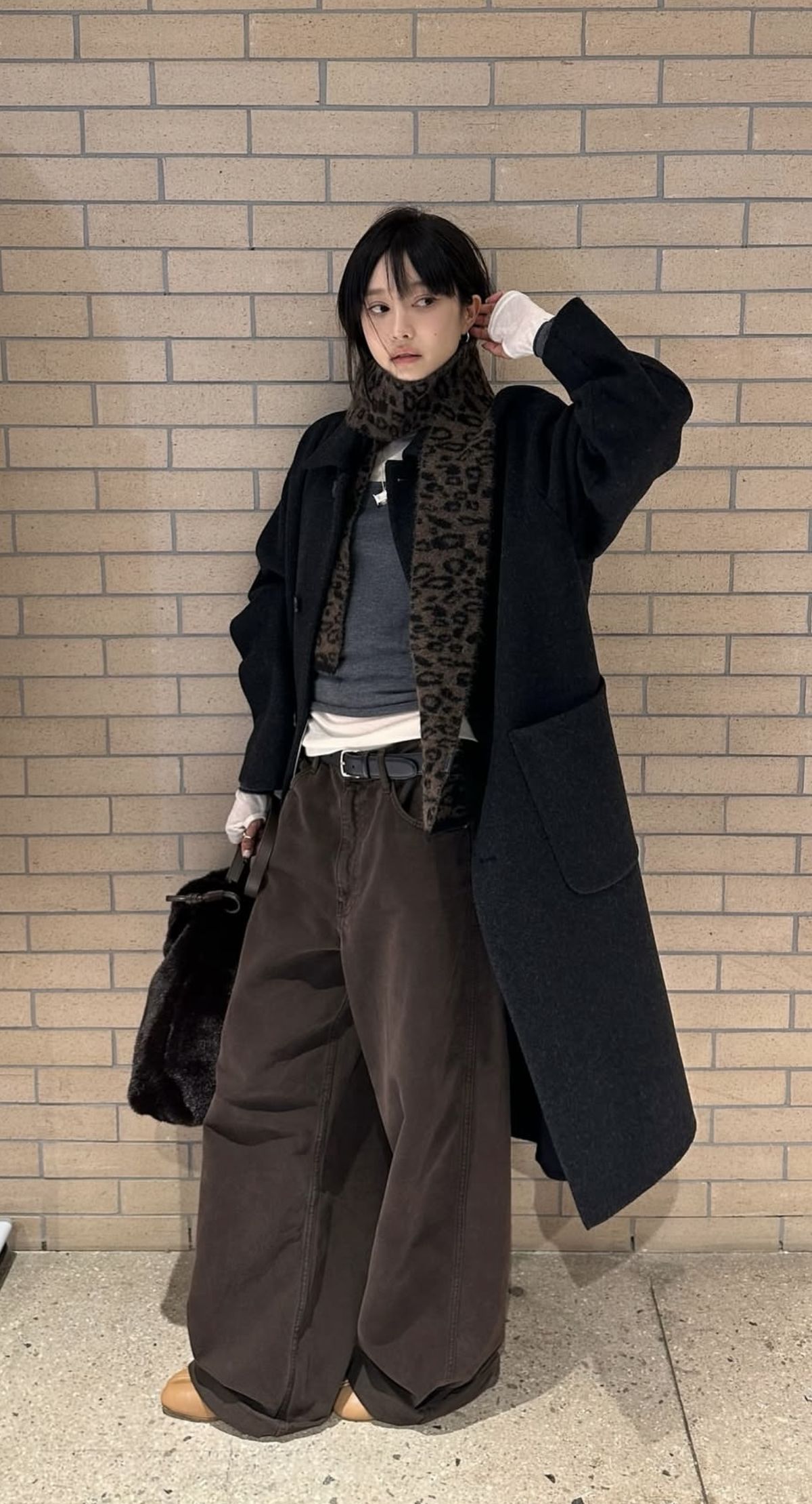 The outfit features an oversized black coat, a gray sweater, and brown wide-leg trousers.
