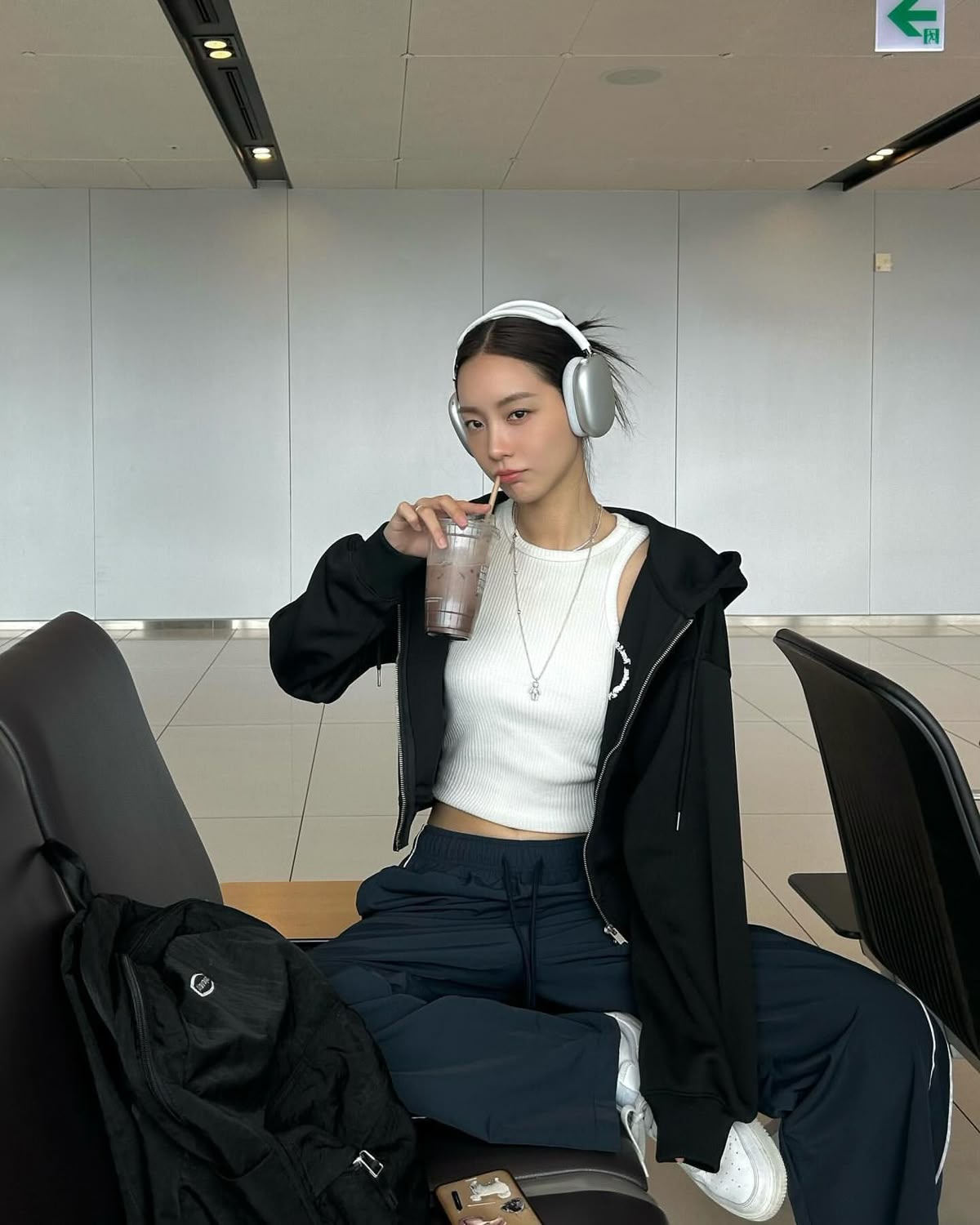 The outfit features a black zip-up hoodie, a white cropped tank top, navy blue joggers, and headphones.