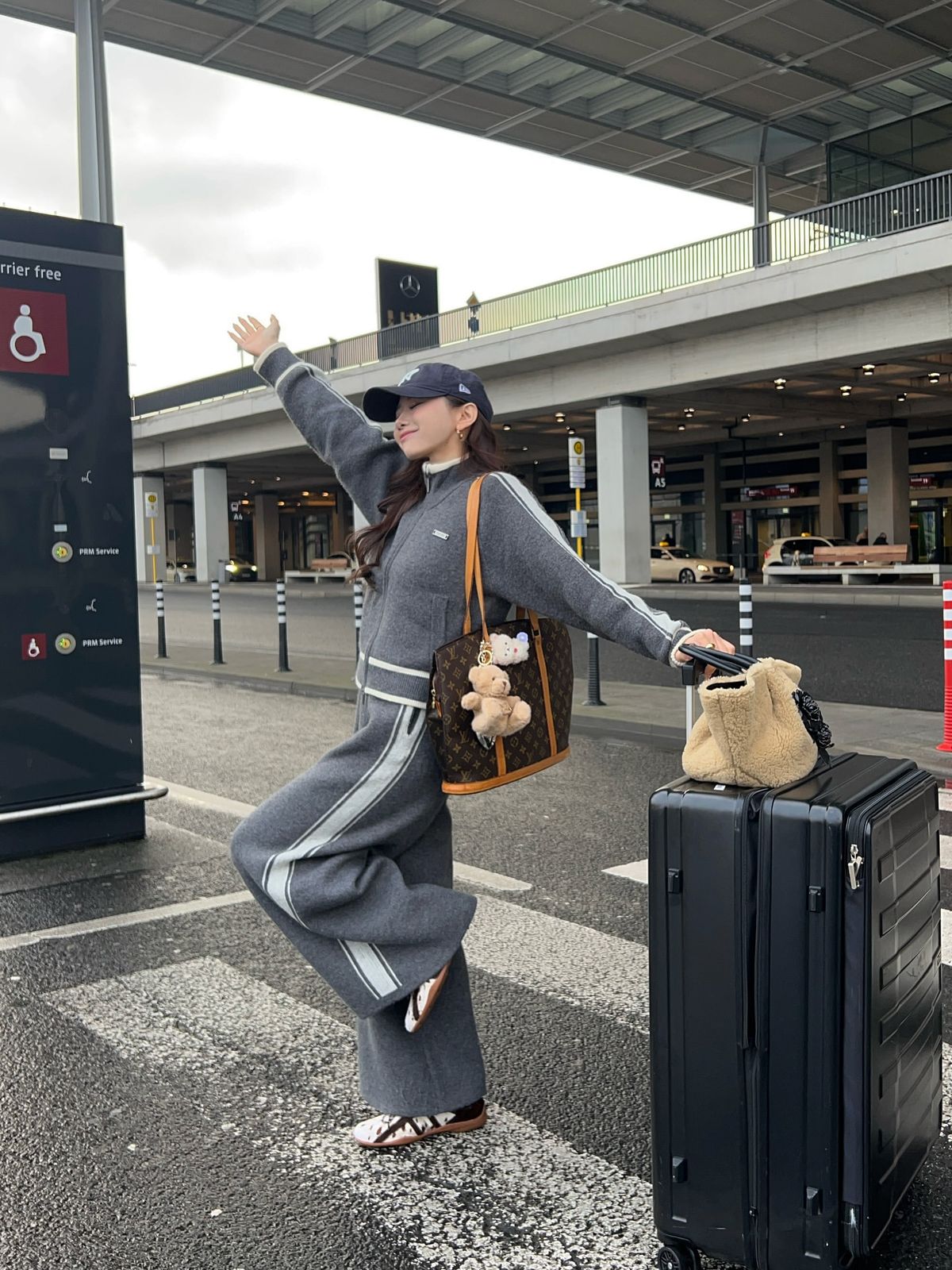 The outfit features a gray zip-up jacket, wide-leg gray pants, a cap, and two bags.