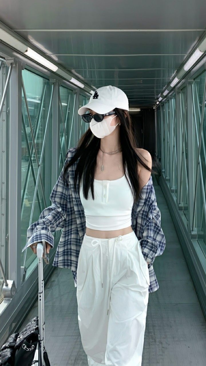 A woman wearing a white crop top, baggy white pants, and a plaid shirt with a baseball cap and sunglasses.